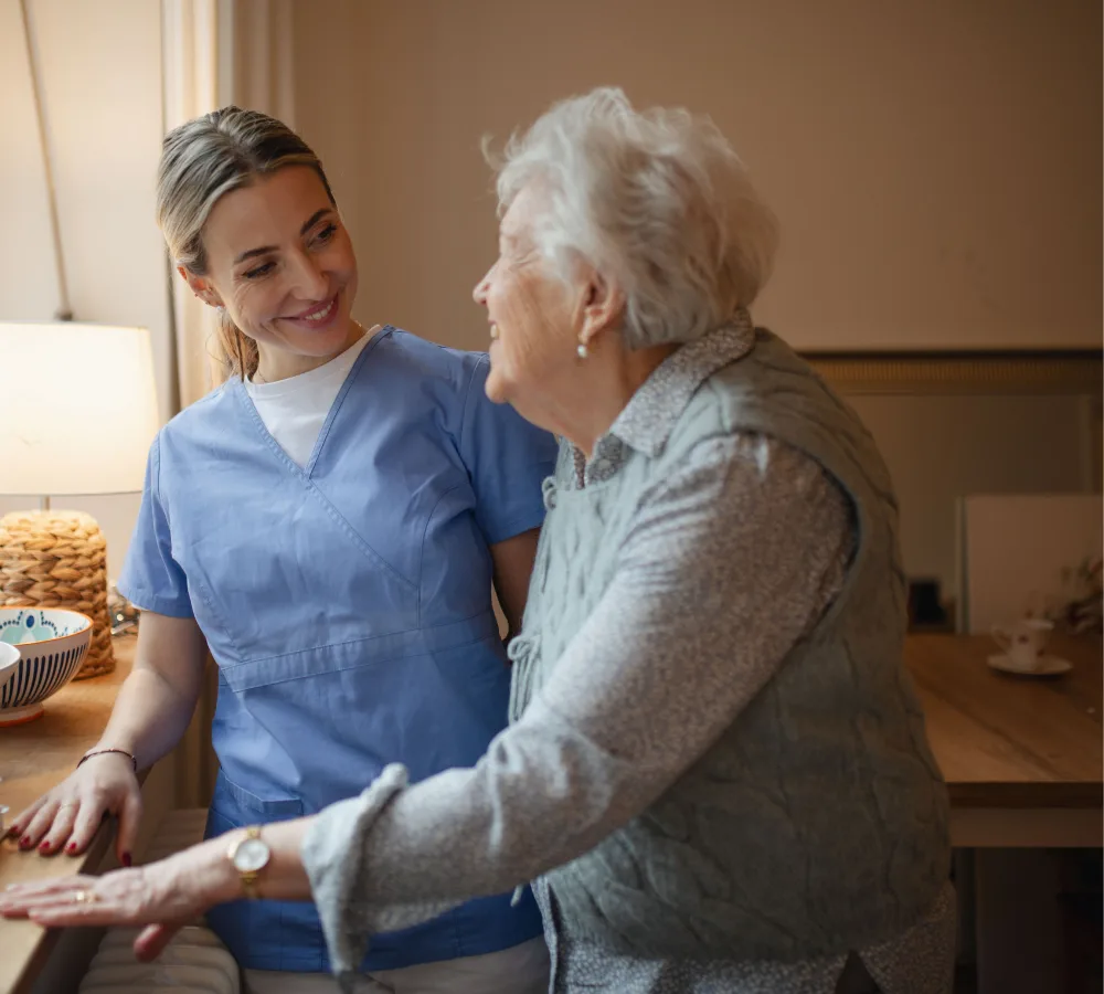 A nurse in blue scrubs smiles warmly at an elderly woman in a cozy room, conveying a caring and supportive atmosphere. A lamp softly illuminates the room.