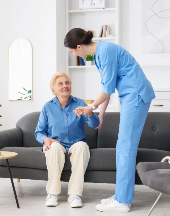 A caregiver in blue scrubs assists an elderly woman seated on a gray sofa. The setting is a bright, modern room, conveying care and support.